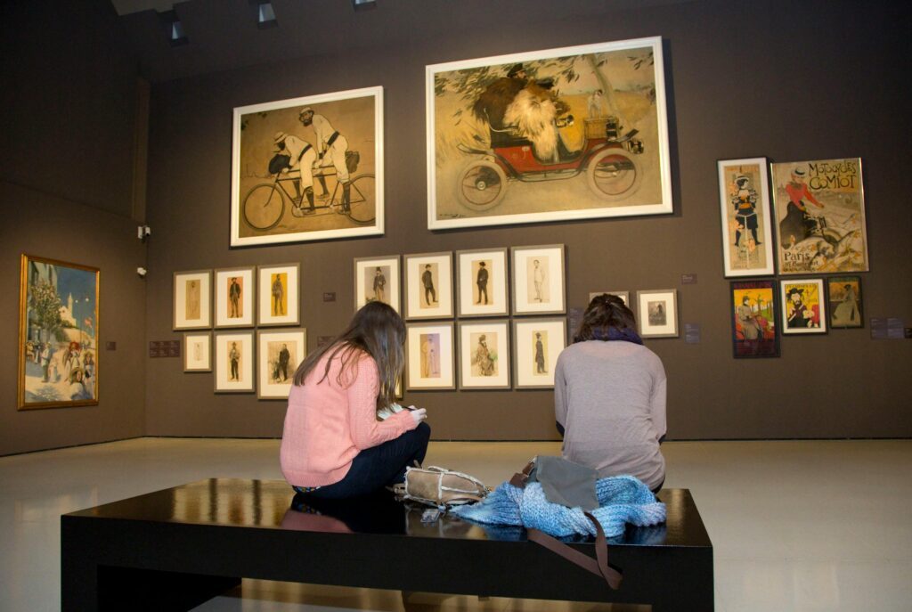 Two people sit on a black bench in an art museum gallery, facing a dark wall filled with framed vintage illustrations and posters, including large paintings of a tandem bicycle and an early automobile displayed above smaller portrait sketches.